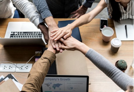 Five aligned team members around a table with arms extended and hands stacked one upon the other